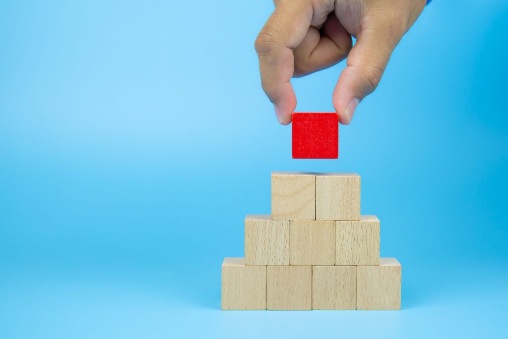 Hand placing red block atop pyramid of wooden blocks symbolizing strong organizational foundation and leadership