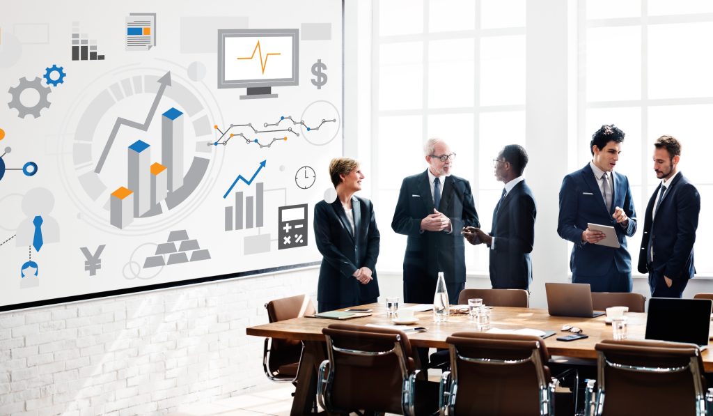 Group of business professionals in discussion with growth charts and business analytics displayed on wall behind them