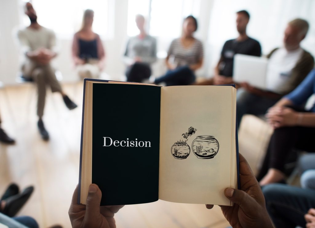 Open book showing "Decision" text with balanced scales icon, held up in front of business team meeting