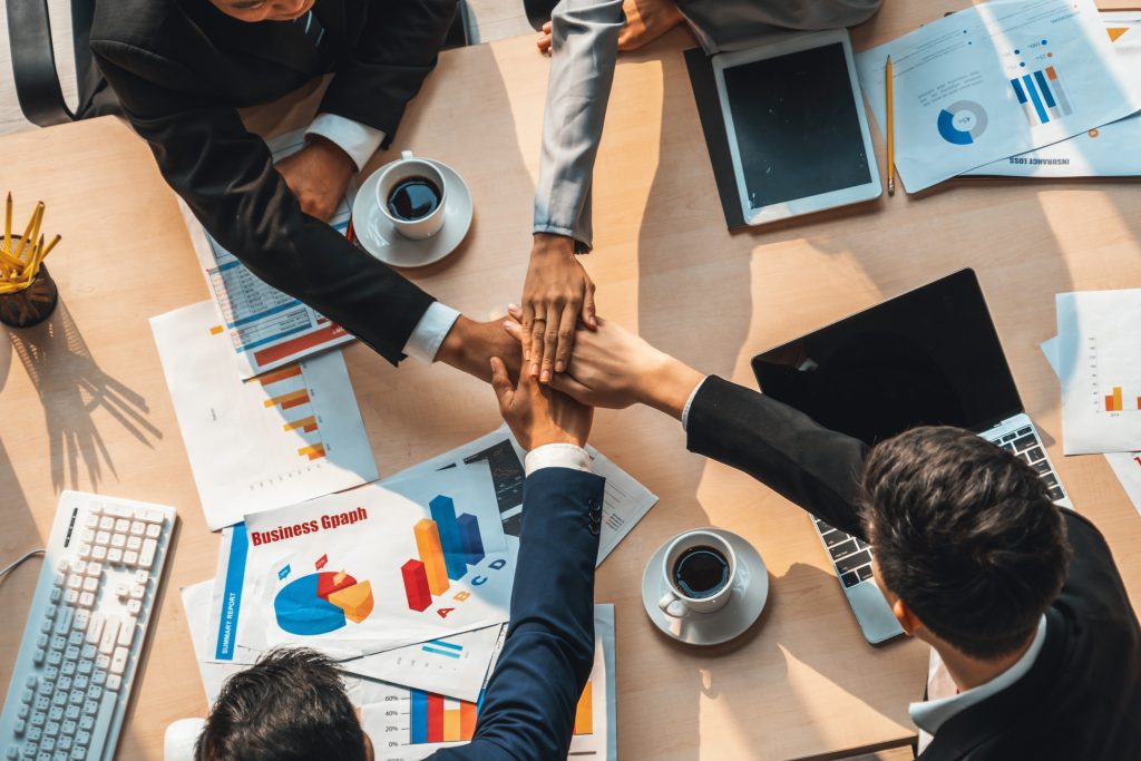 Team members joining hands together over workspace with laptops and charts