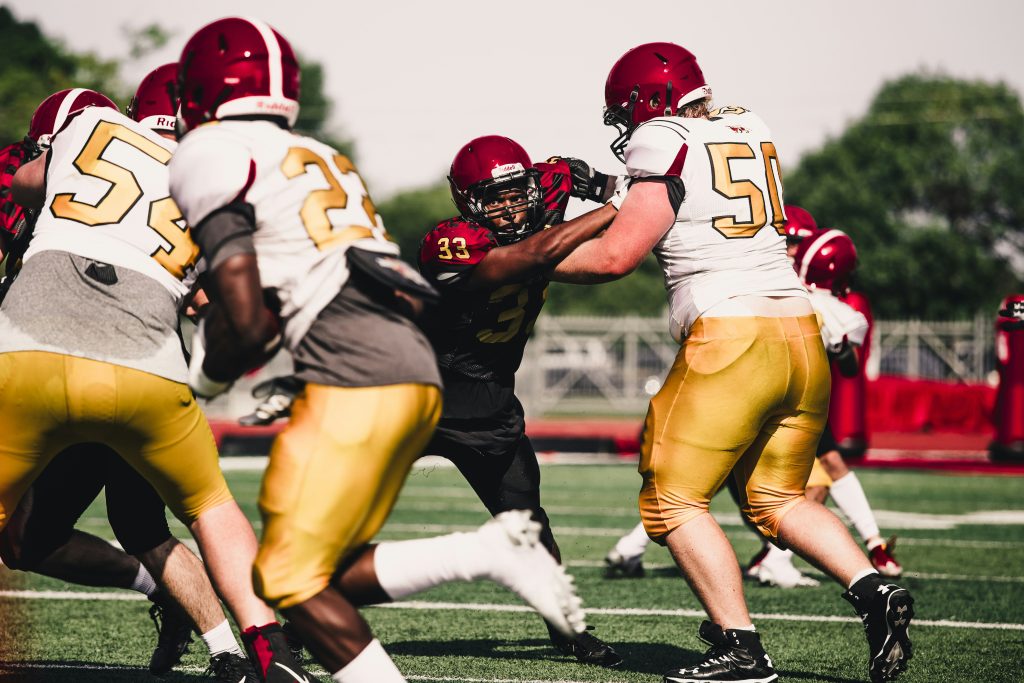 Football player in dark jersey breaking through offensive line in gold jerseys during competitive game