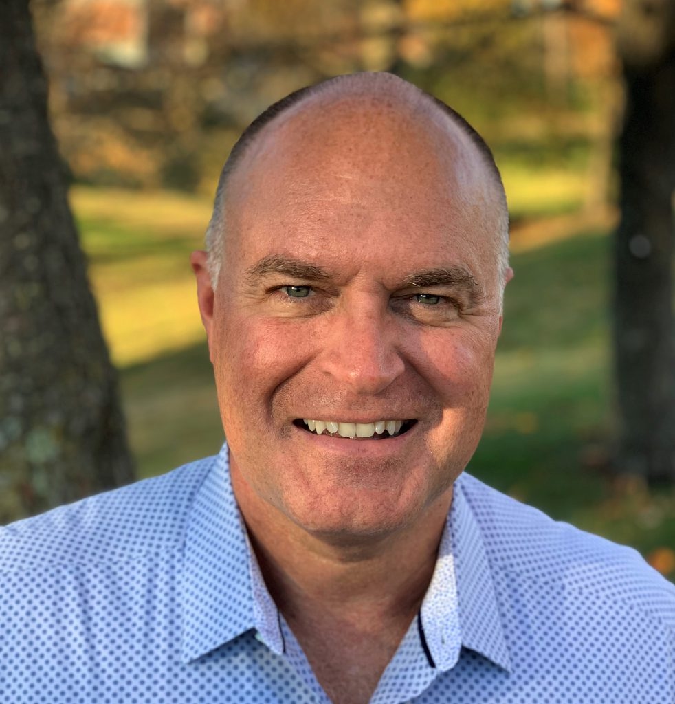 Smiling professional headshot of a man in blue button-up shirt outdoors