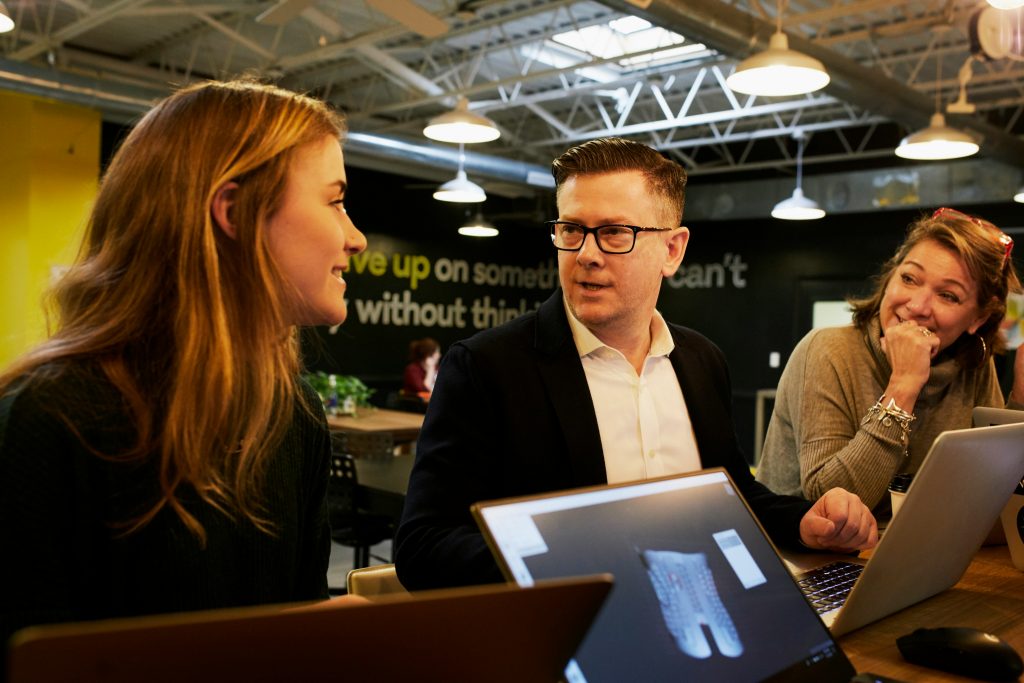 Three professionals in business discussion at a table with laptops, demonstrating sales conversation and client engagement